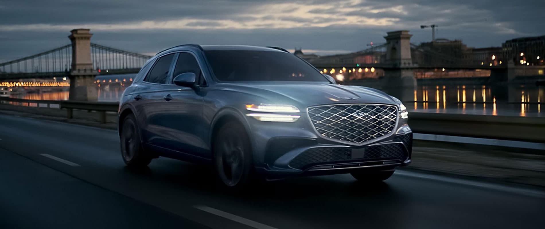 A gray Genesis GV70 driving at night by a river in a city.